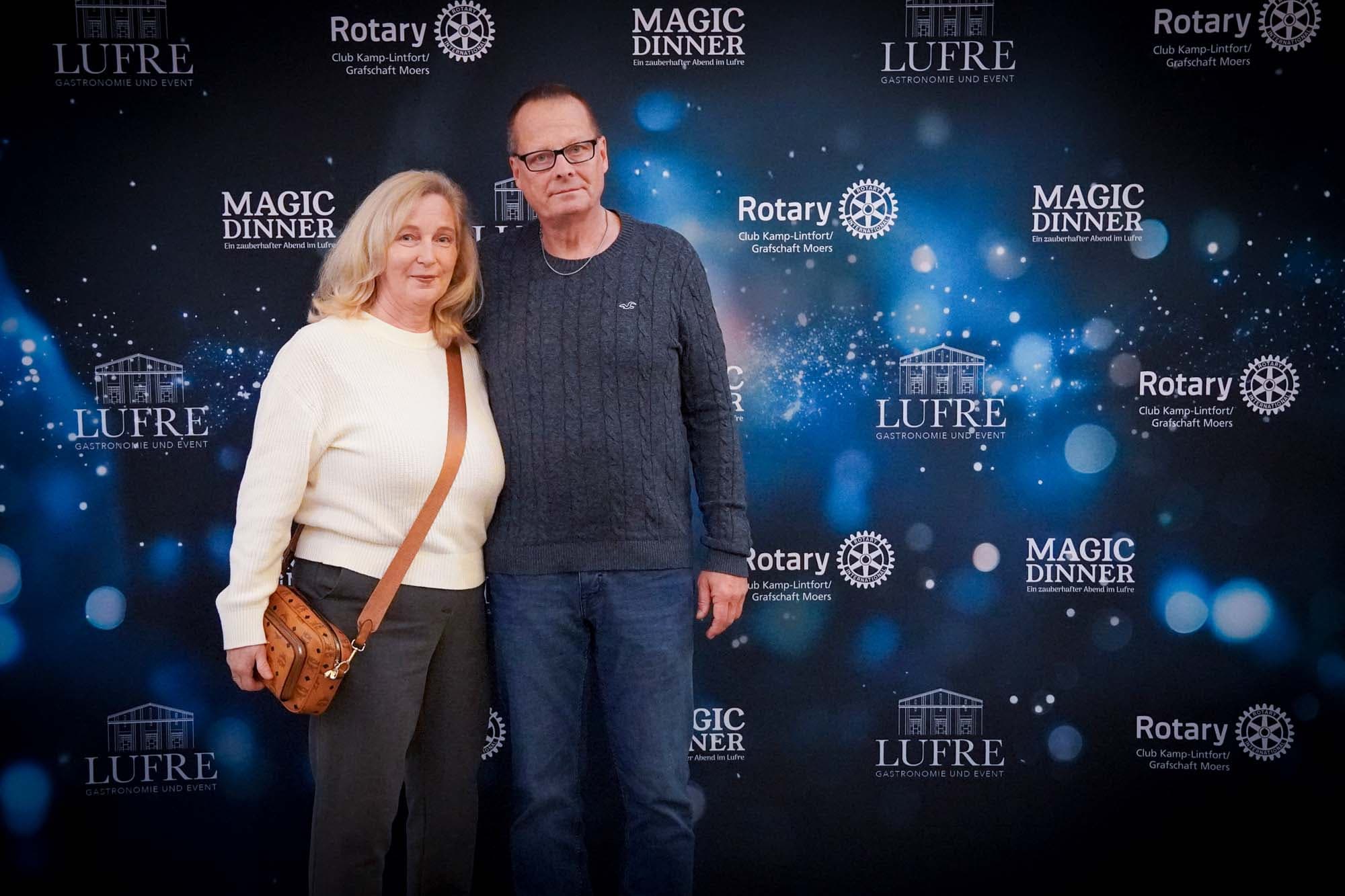 Ein Paar mittleren Alters steht vor der Pressewand, die Frau im weißen Pullover und der Mann in dunklem Hemd mit Brille.