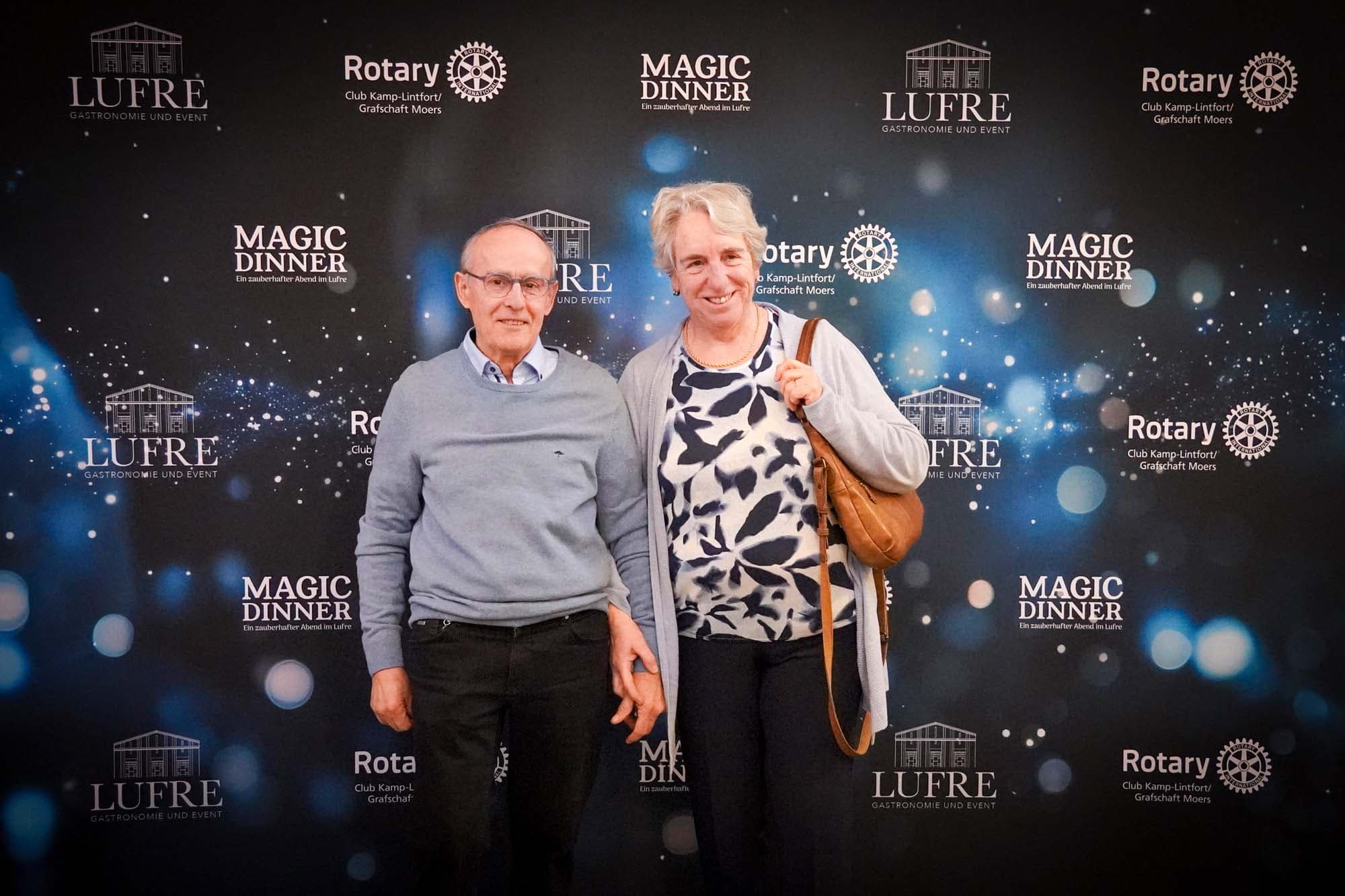 Ein älteres Paar posiert vor der Pressewand, der Mann in grauem Pullover und die Frau mit Umhängetasche in gemustertem Oberteil.