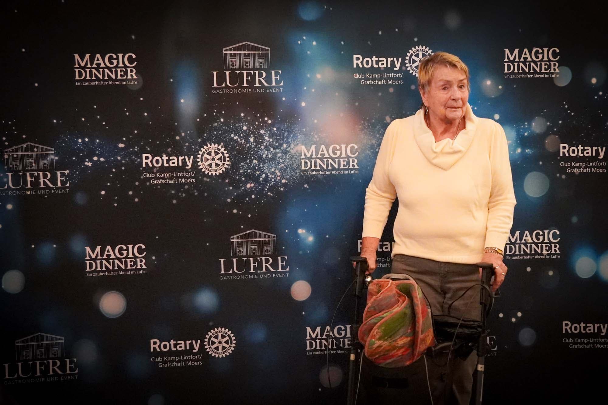 Eine ältere Dame in weißem Pullover steht mit einem Rollator vor der Magic Dinner Pressewand.