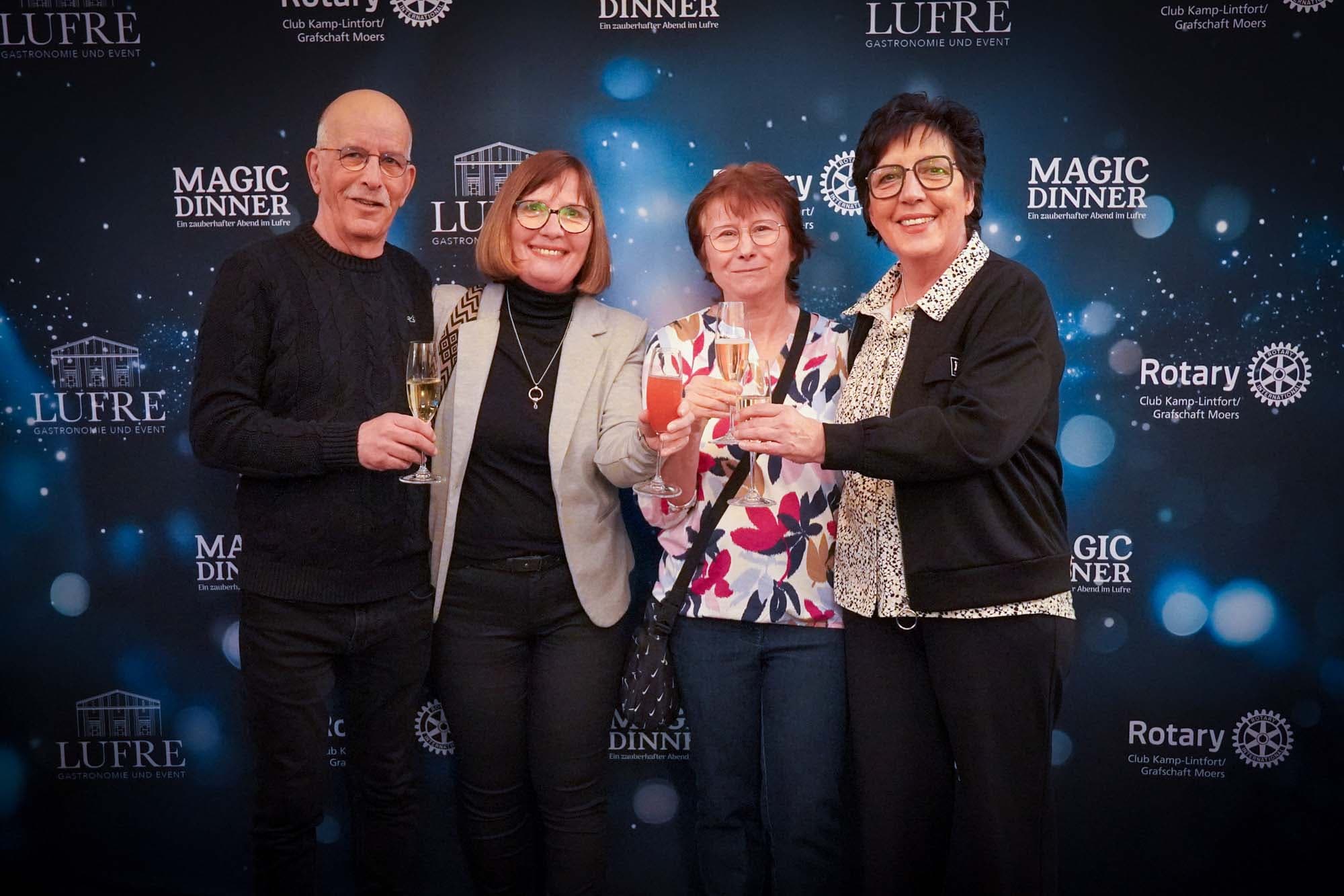 Ein Mann und drei Frauen stoßen fröhlich mit Sektgläsern vor der Magic Dinner Pressewand an.