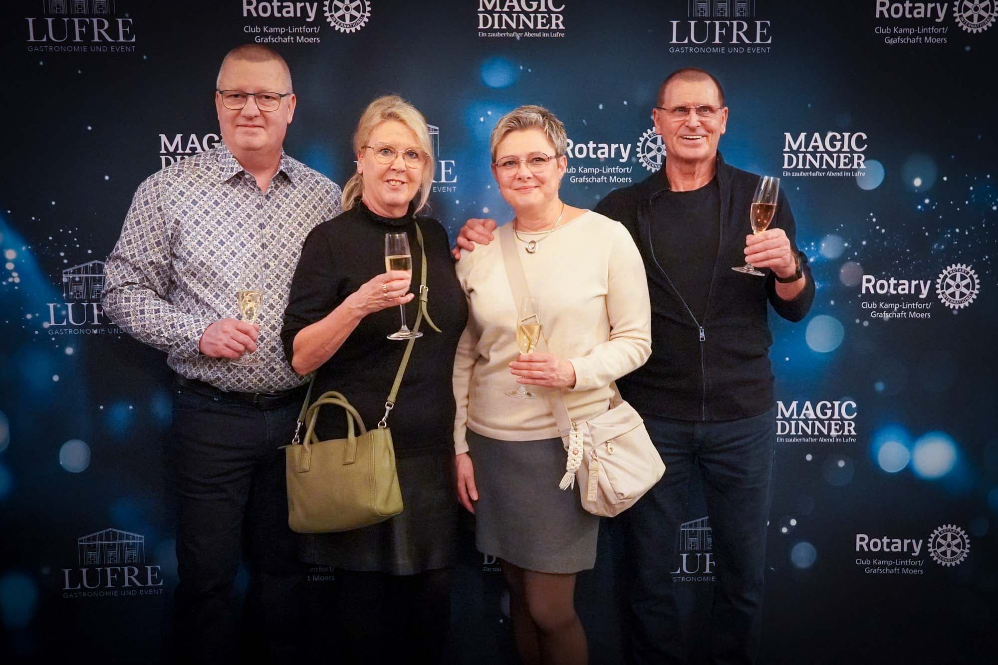 Zwei Paare posieren mit Sektgläsern vor der Magic Dinner Pressewand, die Damen in schwarzer und heller Kleidung, die Herren in gemustertem Hemd und dunklem Outfit.