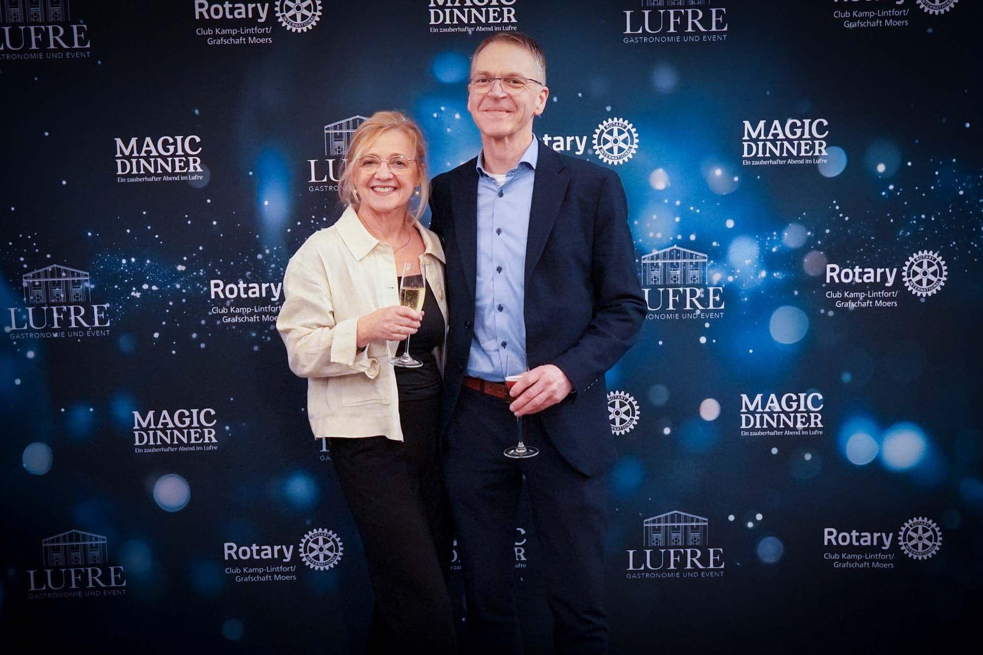 Ein lächelndes Paar vor der Pressewand, sie in heller Jacke mit Sektglas, er im dunkelblauen Sakko mit Brille.