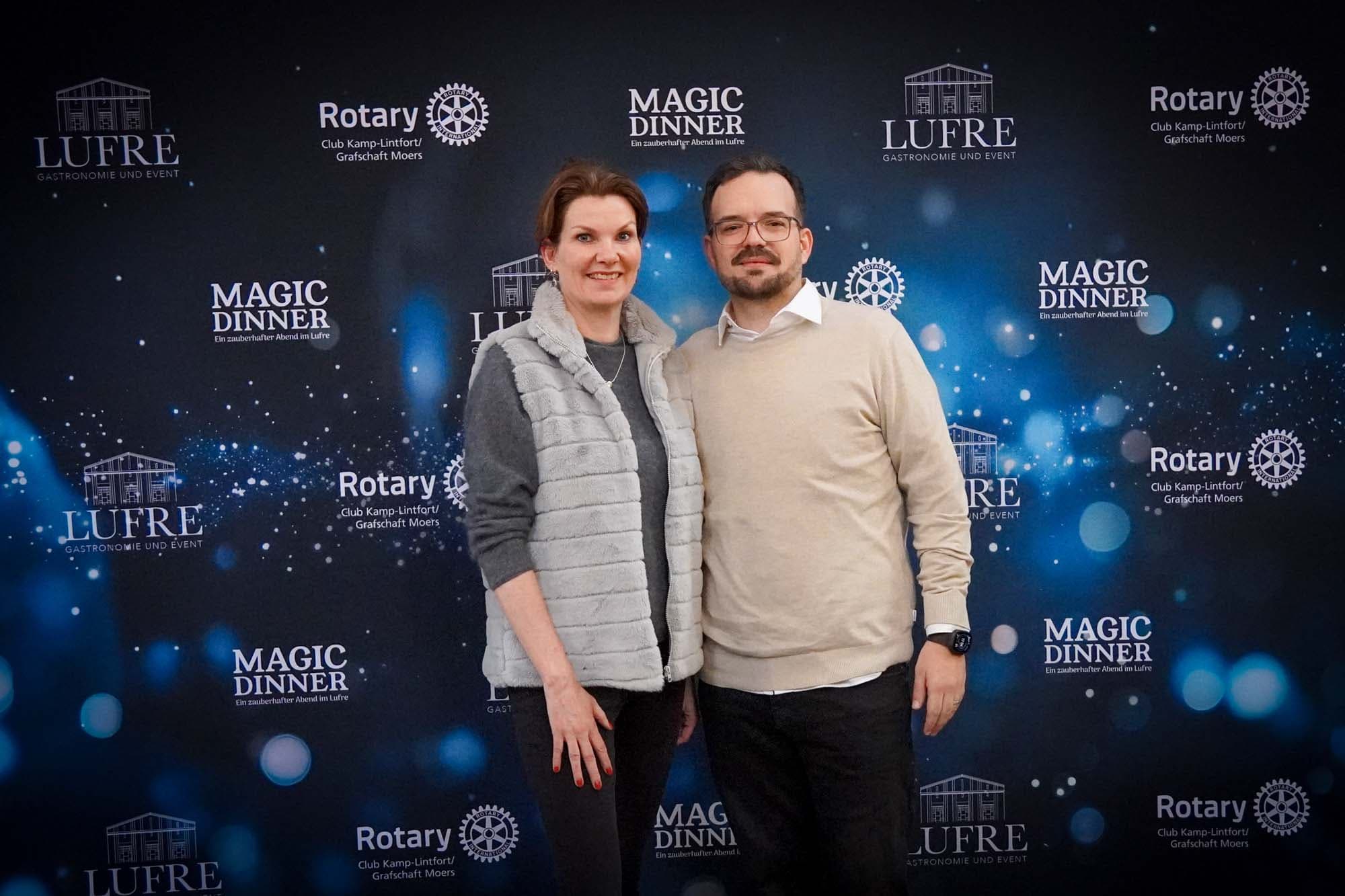 Eine Frau in grauer Jacke und ein Mann mit Brille in beigem Pullover stehen lächelnd vor der Magic Dinner Pressewand.
