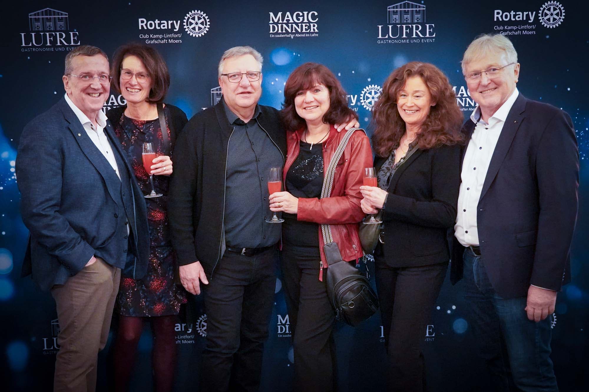 Sechs festlich gekleidete Gäste mit Cocktailgläsern posieren gesellig vor der Pressewand, darunter eine Frau in roter Lederjacke.