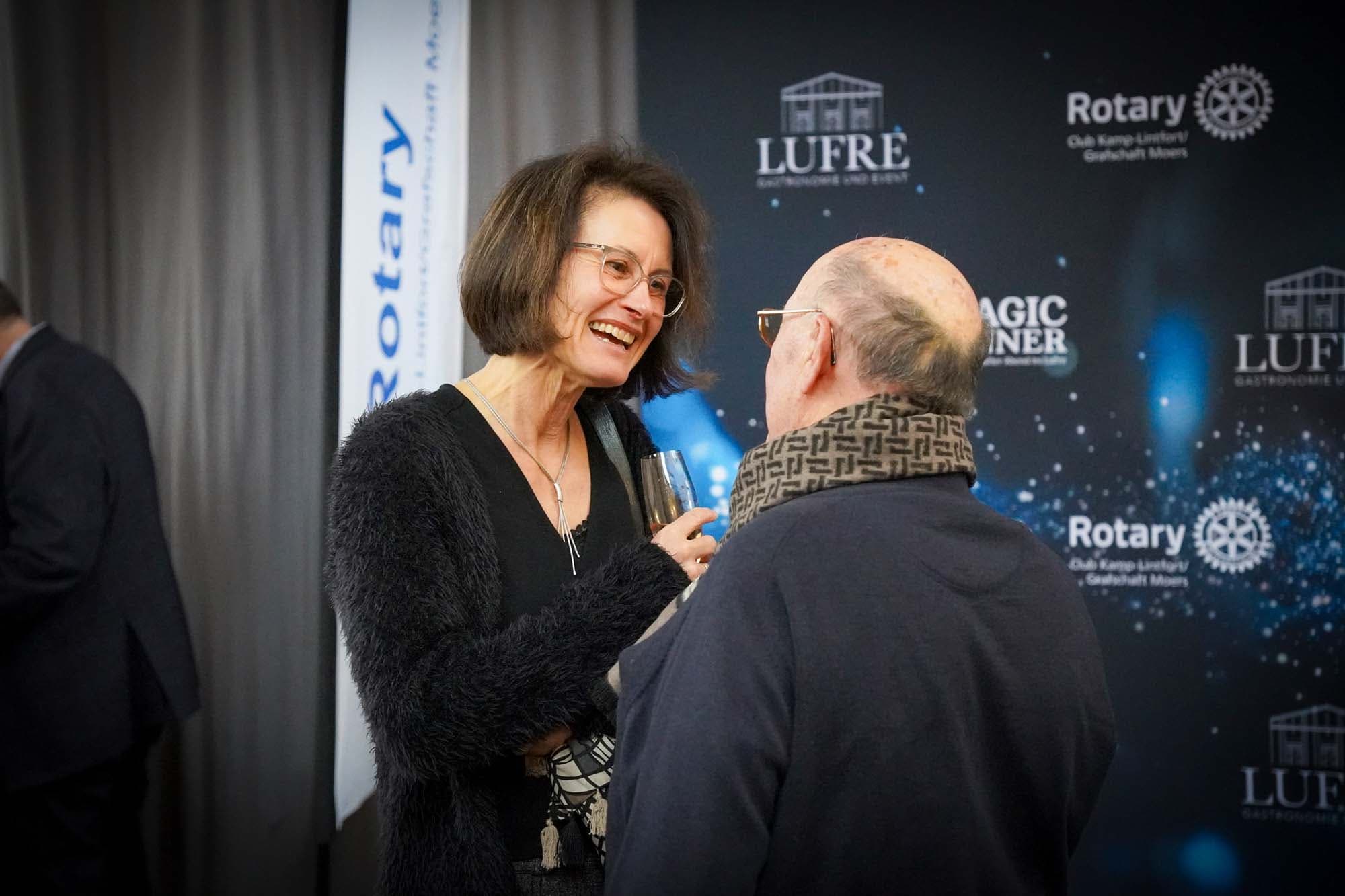 Eine Frau unterhält sich lachend mit einem Gast vor der Sponsorenwand mit Rotary- und Lufre-Logos.