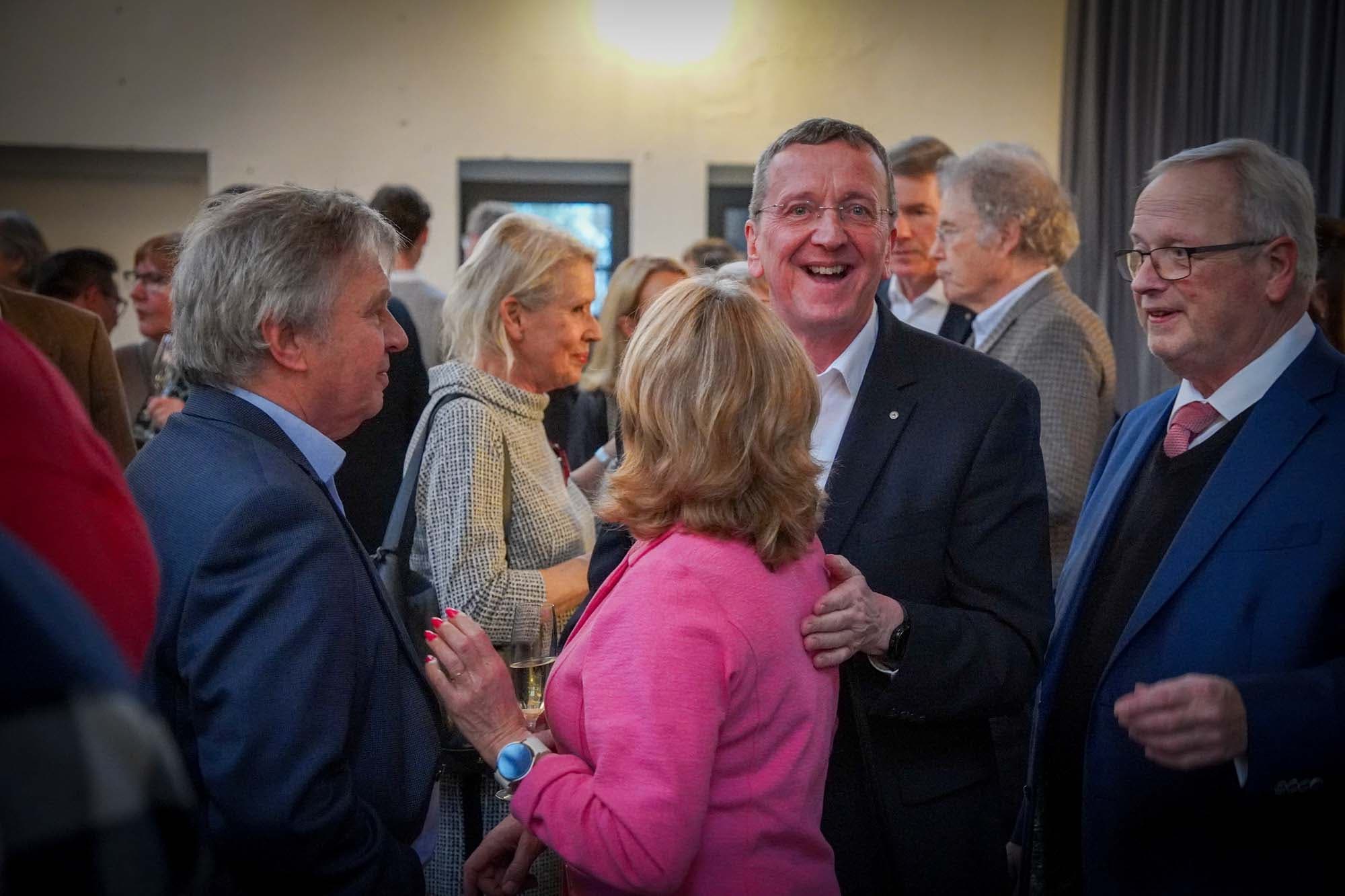 Ein lachender Herr unterhält sich angeregt mit einer Frau im pinkfarbenen Blazer beim Stehempfang.