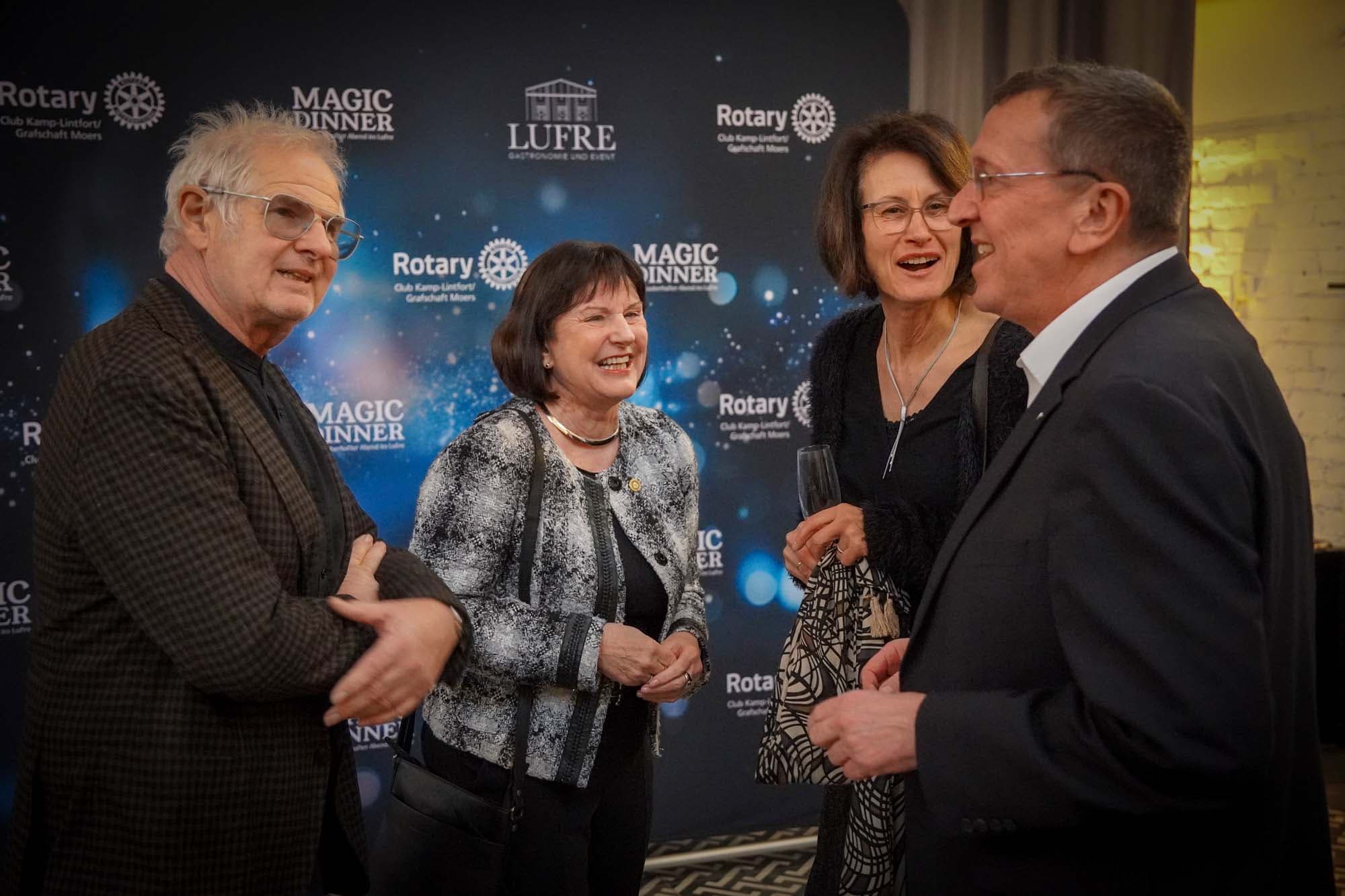 Vier elegant gekleidete Gäste unterhalten sich angeregt vor der Magic-Dinner-Fotowand mit Rotary- und Lufre-Logos.