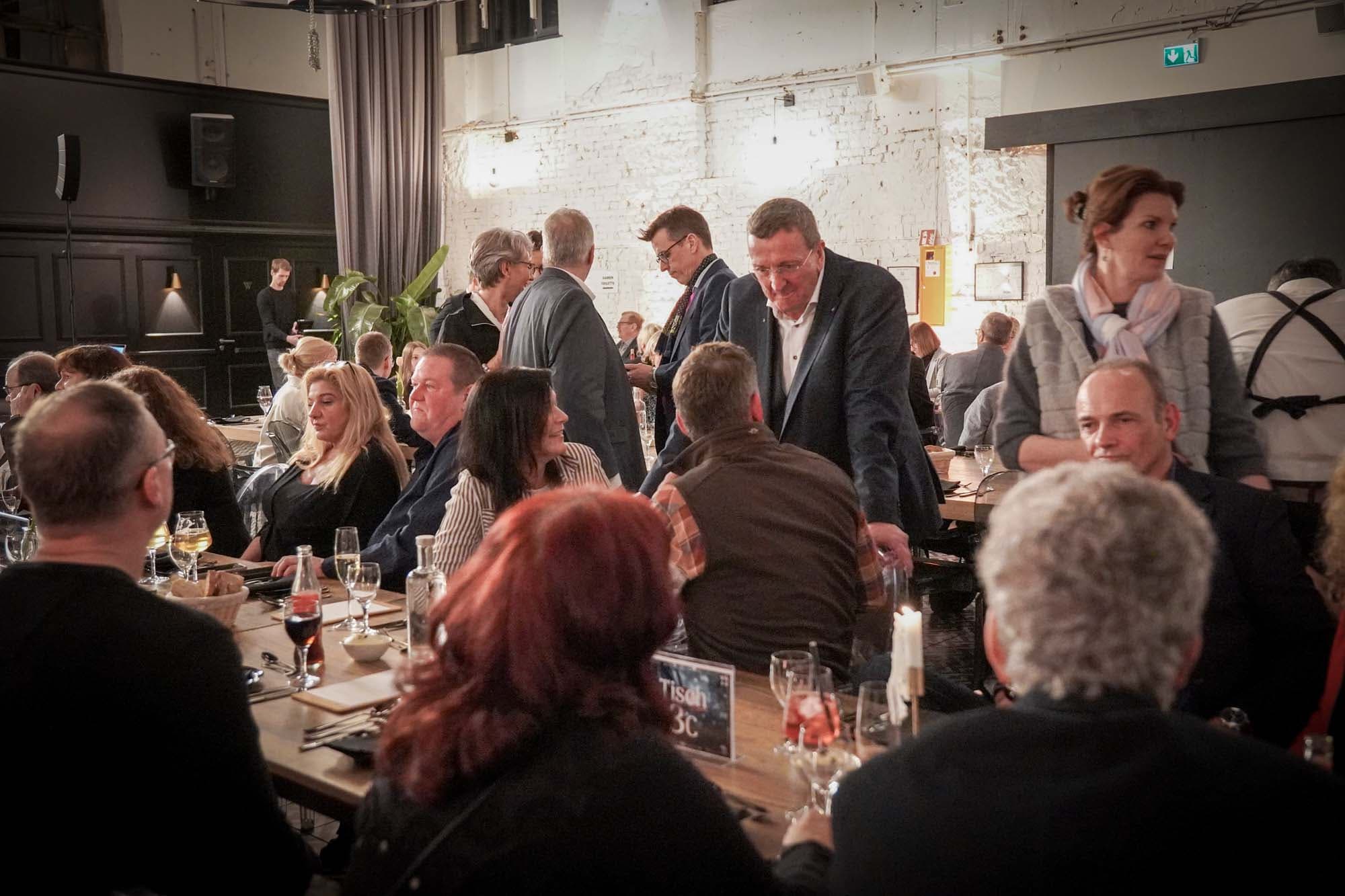 Gäste sitzen an langen Tischen und unterhalten sich angeregt im voll besetzten Saal.